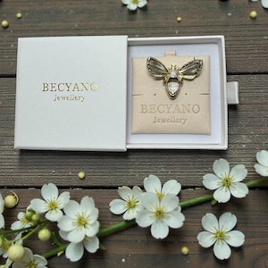 Peut inclure: Un broche en forme d'abeille dorée avec des ailes transparentes et une pierre précieuse centrale, présentée dans un coffret blanc ouvert. Le nom de la marque "BECYANO Jewellery" est imprimé sur le coffret et la carte.