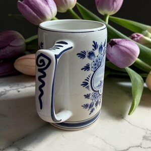 May include: White ceramic mug with blue floral and landscape designs. The mug features a curved handle with a blue spiral design. The mug is decorated with blue floral patterns and a landscape scene. Purple tulips are in the background.