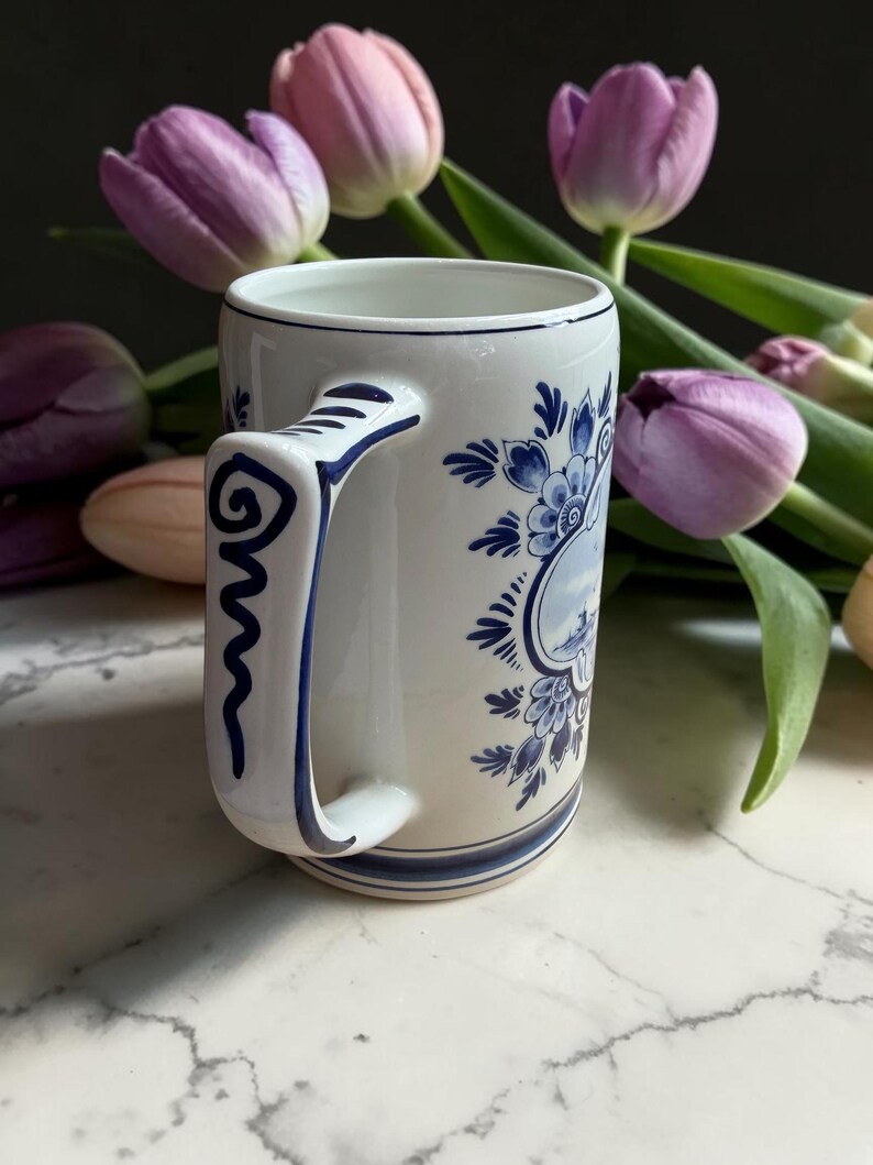 May include: White ceramic mug with blue floral and landscape designs. The mug features a curved handle with a blue spiral design. The mug is decorated with blue floral patterns and a landscape scene. Purple tulips are in the background.