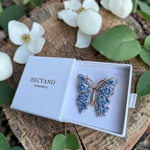 May include: A gold-coloured butterfly brooch with blue floral detailing, displayed in an open white box. The box features the brand name "BECYANO Jewellery". The brooch is set on a wooden surface, surrounded by white flowers and green leaves. A decorative item.