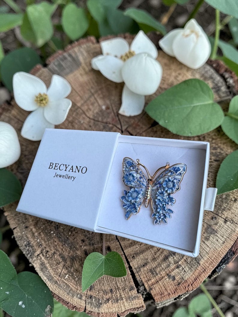 May include: A gold-coloured butterfly brooch with blue floral detailing, displayed in an open white box. The box features the brand name "BECYANO Jewellery". The brooch is set on a wooden surface, surrounded by white flowers and green leaves. A decorative item.