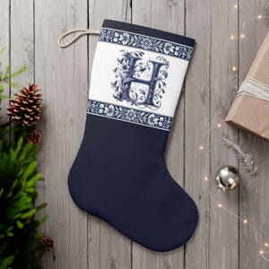 Op de afbeelding: Marineblauwe kerstsok met een wit paneel met een bloemenpatroon en de letter "H" in blauw. De sok heeft een lus om op te hangen en ligt op een houten achtergrond met feestelijke decoraties.