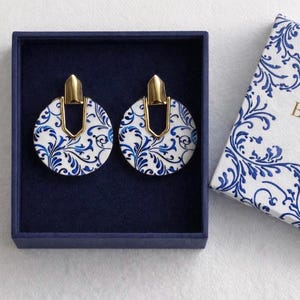 Può includere: Un paio di orecchini rotondi con un motivo floreale bianco e blu, in una scatola blu navy. Gli orecchini hanno dettagli in metallo color oro. Una scatola con motivo coordinato è parzialmente visibile.