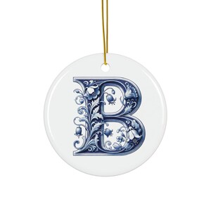 Op de afbeelding: Een wit keramiek ornament met een gouden ophangtouwtje. Het ornament heeft een blauw bloemmotief in de vorm van de letter "B". Het bloemmotief is gedetailleerd met bladeren en klokvormige bloemen.