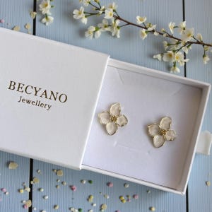 Può includere: Orecchini a forma di fiore in un cofanetto bianco. Gli orecchini hanno una montatura color oro e un centro bianco traslucido con una piccola perla dorata. La scatola bianca riporta la scritta "BECYANO Jewellery".
