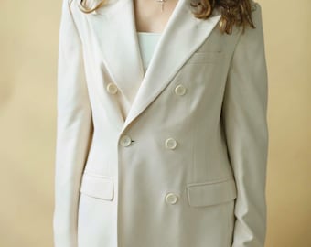 Puede incluir: Una americana de doble botonadura en color crema con solapas de muesca, mangas largas y cuatro botones redondos en la parte delantera. La chaqueta incluye dos bolsillos laterales con solapa y un bolsillo en el pecho, sobre un top blanco.