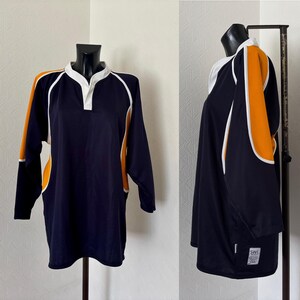 Könnte beinhalten: Marineblaues Rugby-Shirt mit langen Ärmeln und weißen und orangefarbenen Akzenten. Das Shirt hat einen weißen Kragen und eine Knopfleiste. Orangefarbene Einsätze mit weißer Paspelierung an den Ärmeln. Auf einer schwarzen Schaufensterpuppe.