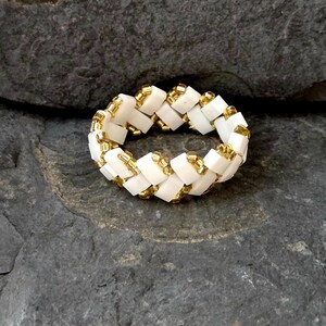 Op de afbeelding: Een witte en gouden kralenring. De ring is gemaakt van kleine, vierkante witte kralen en goudkleurige kralen. De kralen zijn in een geweven patroon gerangschikt. De ring ligt op een donkergrijze, getextureerde achtergrond.