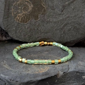 Könnte beinhalten: Ein Armband mit hellgrünen rechteckigen Perlen und goldfarbenen zylindrischen Perlen. Das Armband ist auf einer goldfarbenen Kordel aufgefädelt. Das Armband wird auf einer dunkelgrauen Steinoberfläche gezeigt.