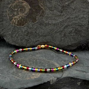 Könnte beinhalten: Ein mehrfarbiges Perlenarmband mit einem goldenen Verschluss. Das Armband besteht aus kleinen, runden Perlen in Regenbogenfarben, darunter Rot, Gelb, Grün, Blau und Lila. Das Armband wird auf einer dunkelgrauen, strukturierten Oberfläche präsentiert.