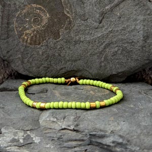 Könnte beinhalten: Ein Armband mit kleinen, hellgrünen Perlen und goldfarbenen zylindrischen Perlen. Das Armband ist auf einem flexiblen Band aufgefädelt und wird auf einer grauen Steinoberfläche präsentiert. Es ist ein Schmuckstück.