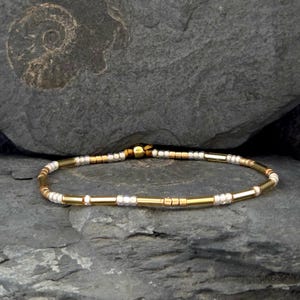 Könnte beinhalten: Ein filigranes Armband mit einer Mischung aus goldenen und silbernen Perlen und zylindrischen Stäben. Das Armband ist auf einem dünnen, flexiblen Band aufgefädelt und liegt auf einer dunkelgrauen Steinoberfläche. Das Design des Armbands ist schlicht und elegant.