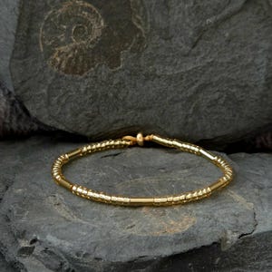 Könnte beinhalten: Ein goldfarbenes Armband mit Perlenmuster. Das Armband liegt auf einer dunkelgrauen Steinoberfläche. Das Armband besteht aus kleinen, zylindrischen Perlen und längeren, glatten Segmenten.