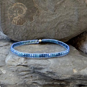 Könnte beinhalten: Ein zartes blaues und silbernes Perlenarmband mit einem Magnetverschluss. Das Armband besteht aus kleinen, quadratischen Perlen, die in einem abgestuften Muster angeordnet sind, wobei die dunkelsten blauen Perlen in der Mitte und die hellsten blauen Perlen an den Rändern liegen.