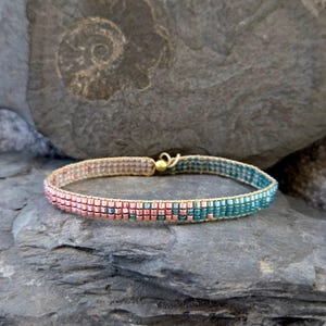 Könnte beinhalten: Ein zartes Armband mit Perlen in einem zweifarbigen Design. Das Armband besteht aus kleinen, quadratischen Perlen in Rosétönen und Türkis, die durch einen dünnen goldenen Draht getrennt sind. Das Armband wird mit einem goldenen Verschluss geschlossen.