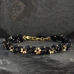 Könnte beinhalten: Ein schwarzes Perlenarmband mit goldfarbenen Blumenakzenten. Das Armband hat einen goldenen Verschluss und wird auf einer dunkelgrauen Steinoberfläche präsentiert. Die floralen Details verleihen dem Schmuck einen Hauch von Eleganz.