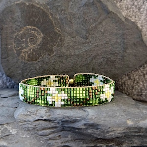 Könnte beinhalten: Ein grünes und weißes Armband mit Blumenmuster. Das Armband ist aus kleinen, quadratischen Perlen gefertigt und hat einen goldenen Verschluss.