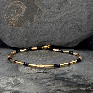 Könnte beinhalten: Ein gold-schwarzes Perlenarmband. Das Armband hat abwechselnd goldfarbene zylindrische Perlen und kleine schwarze Perlen. Das Armband wird auf einer grauen Steinoberfläche präsentiert.