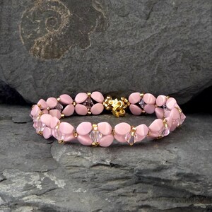 Könnte beinhalten: Ein rosa Perlenarmband mit goldenen Akzenten und einem goldenen Verschluss. Das Armband hat ovale rosa Perlen und klare Kristallperlen, die ein zartes Design ergeben. Das Armband wird auf einer grauen Steinoberfläche präsentiert.