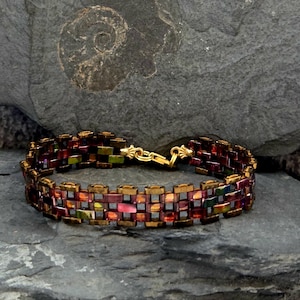 Könnte beinhalten: Ein Perlenarmband mit einem goldenen Verschluss. Das Armband besteht aus kleinen, rechteckigen Perlen in Rot-, Gold-, Grün- und Lilatönen. Das Armband wird auf einer strukturierten, grauen Oberfläche präsentiert.