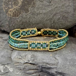 Könnte beinhalten: Ein mehrreihiges Perlenarmband mit einem Farbverlauf aus grünen und goldenen Perlen. Das Armband ist aus kleinen, quadratischen Perlen gefertigt und hat ein schlichtes, elegantes Design.