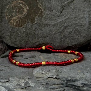 Könnte beinhalten: Rotes Perlenarmband mit goldfarbenen Akzenten. Das Armband besteht aus kleinen, roten Perlen und goldfarbenen zylindrischen Perlen. Es liegt auf einer grauen Steinoberfläche. Die Länge beträgt ca. 18 cm.