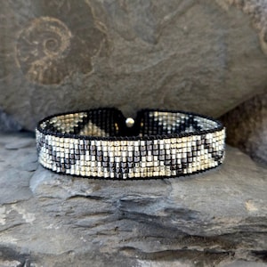 Könnte beinhalten: Ein schwarzes und silbernes Perlenarmband mit geometrischem Muster. Das Armband besteht aus kleinen, quadratischen Perlen, die in einem sich wiederholenden Muster angeordnet sind.