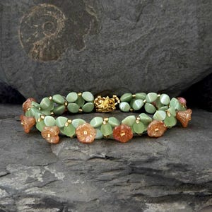 Könnte beinhalten: Ein doppelreihiges Armband mit hellgrünen ovalen Perlen und kleinen orangefarbenen blütenförmigen Perlen. Das Armband hat einen goldfarbenen Verschluss und wird auf einer dunkelgrauen Steinoberfläche präsentiert. Ein einzigartiges Schmuckstück.