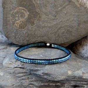 Könnte beinhalten: Ein blaues und schwarzes Perlenarmband mit einem silbernen Verschluss. Das Armband besteht aus kleinen, quadratischen Perlen, die in einem Farbverlauf angeordnet sind.