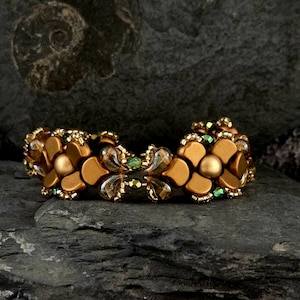 Könnte beinhalten: Ein handgefertigtes Armband mit floralem Design. Das Armband besteht aus goldfarbenen Perlen und Akzenten mit einem zentralen Blumendesign. Kleine grüne Perlen setzen Farbakzente. Das Armband wird auf einer dunklen, strukturierten Oberfläche präsentiert.