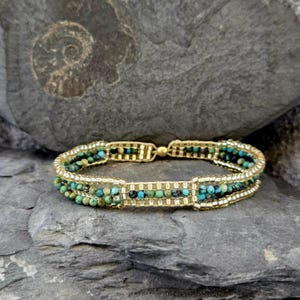 May include: A gold beaded bracelet with three rows of turquoise and gold beads. The bracelet is on a gray and green background.