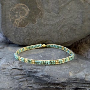 Könnte beinhalten: Ein zartes Armband aus goldenen und türkisfarbenen Perlen mit einem Magnetverschluss. Das Armband ist auf einem grauen Steinuntergrund abgebildet.