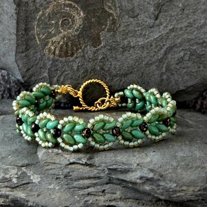 Könnte beinhalten: Ein Armband mit Perlen, das ein sich wiederholendes Muster aus türkis- und schwarzen Perlen aufweist, akzentuiert mit weißen Perlen. Das Armband wird mit einem goldfarbenen Verschluss geschlossen. Das Schmuckstück wird auf einer grauen, strukturierten Oberfläche präsentiert.