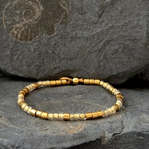Könnte beinhalten: Ein Armband mit einer Mischung aus goldenen und klaren Perlen. Das Armband ist an einem beigefarbenen Kordelband befestigt und wird auf einer grauen Steinoberfläche präsentiert. Die Perlen sind klein und gleichmäßig verteilt.