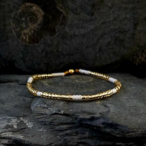 Könnte beinhalten: Ein goldfarbenes Perlenarmband mit weißen Akzenten liegt auf einer dunkelgrauen Steinoberfläche. Das Armband besteht aus kleinen, gleichmäßig verteilten Perlen in Gold und Weiß, die zu einem flexiblen Band aufgereiht sind. Das Armband ist ein zartes Schmuckstück.