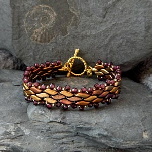 Könnte beinhalten: Ein gold- und bronzefarbenes Armband mit Knebelverschluss. Das Armband hat ein gewebtes Design mit kleinen, runden, dunkelroten Perlen. Das Armband liegt auf einer grauen Steinoberfläche.