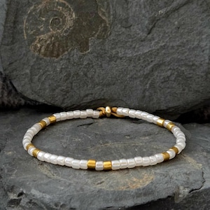 Könnte beinhalten: Ein zartes Armband mit kleinen, weißen Perlen, die mit goldfarbenen Akzenten durchsetzt sind. Das Armband ist auf einer dünnen Schnur aufgefädelt und wird vor einem dunklen, strukturierten Steinhintergrund präsentiert. Das Design des Armbands ist schlicht und elegant.