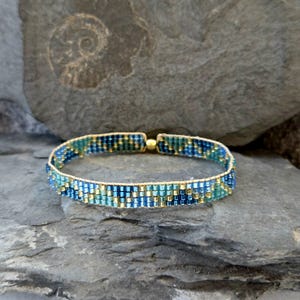 Könnte beinhalten: Ein blaues und goldenes Armband mit geometrischem Muster. Das Armband besteht aus kleinen, quadratischen Perlen und hat einen goldenen Verschluss.