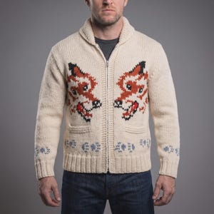 May include: A cream-colored knit zip-up cardigan featuring a shawl collar and two front pockets. The sweater displays a prominent intarsia pattern of two fox faces in orange, black, and white, with small blue geometric motifs along the hem and cuffs.