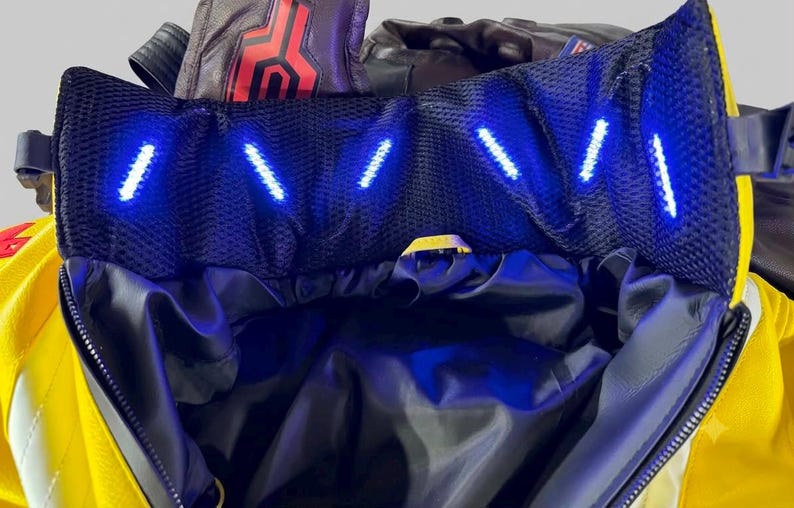 May include: A bright yellow and black jacket featuring a high collar lined with black mesh fabric. The collar contains a series of small, bright blue LED lights arranged in a row. The jacket has a dark interior lining and a front zipper closure.