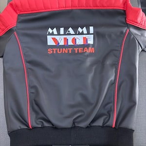 Könnte beinhalten: Eine schwarz-rote Jacke mit gestepptem Schulterdesign und roten Paspeln. Auf der Rückseite befindet sich der weiß-rote Schriftzug MIAMI VICE STUNT TEAM. Die Jacke hat einen schwarzen Rippsaum und einen durchgehenden Frontreißverschluss.