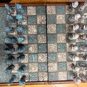Op de afbeelding: Een gedetailleerd schaakspel met een turquoise en bruin kleurenschema. Het bord heeft een turquoise en wit geblokt patroon met gebeeldhouwde Maya-geïnspireerde ontwerpen. De schaakstukken zijn turquoise en wit, met gedetailleerde gravures van Maya-figuren en schedels.
