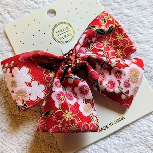 Puede incluir: Lazo de tela rojo y blanco con estampado floral dorado y un centro brillante. El lazo está en una tarjeta blanca con las palabras "Hand Made" y "Made in China".