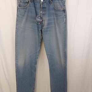Könnte beinhalten: Hellblaue Jeans aus Denim mit verwaschener Optik. Die Jeans haben ein klassisches Fünf-Taschen-Design, einen Knopfverschluss und ein gerades Bein. Ein Etikett mit dem Text "RETRO JEANS" ist angebracht. Die Jeans hängen an einem schwarzen Kleiderbügel.