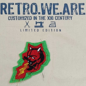 Puede incluir: Un parche de tela con la cara de un diablo rojo, cuernos blancos y un rayo amarillo, delineado en verde. El texto azul en la parte superior dice "RETRO.WE.ARE" y "CUSTOMIZED IN THE XXI CENTURY". Debajo, se lee "LIMITED EDITION".