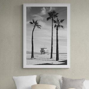 Puede incluir: Una fotograf&iacute;a en blanco y negro de tres palmeras en una playa con una caseta de salvavidas en el fondo. La fotograf&iacute;a est&aacute; enmarcada y colgada en una pared sobre una cama blanca con almohadas.