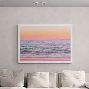 May include: A white sofa with pillows sits in a living room with a white rug and a black woven coffee table. A framed photograph of a beach scene with pink and blue hues hangs on the wall above the sofa.