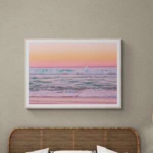 May include: A wooden bed frame with a woven headboard and a white bedspread. The bed is decorated with white and black patterned pillows. A framed photograph of a beach scene with a pink and blue sunset hangs on the wall above the bed.