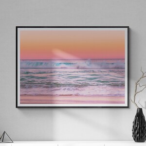 May include: A framed photograph of a pink and blue sunset over a beach with waves crashing on the shore. The photo is hanging on a white wall above a white cabinet with black legs.