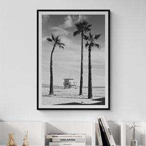 May include: A black and white photograph of three palm trees on a beach with a lifeguard stand in the distance. The photograph is framed in a black frame and is hanging on a white wall above a white bookshelf with books and decorative objects.